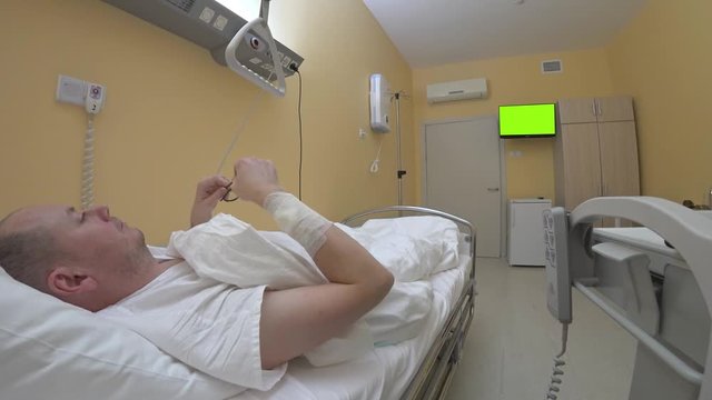 Patient lying in bed puts on glasses and sends a message on his smartphone to his loved ones. Hospital ward lonely patient after surgery chromakey green TV.