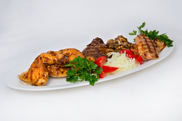 Chicken legs, young veal, slices of young lamb and the back of a grilled piglet on a white oval plate with onions, tomatoes and parsley