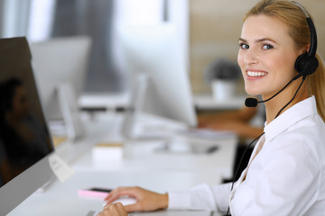 Blonde business woman using headset for communication and consulting people at customer service office. Call center. Group of operators at work at the background