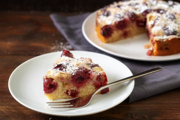 Slice of a juicy cherry cake with icing sugar on a white plate and a dark rustic wooden table