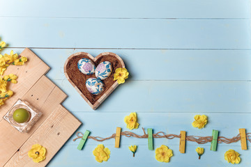 Heart shaped box with decorated Easter eggs, yellow flowers, burning candle and brown boards on wooden blue background. Spring and Easter concept. Top view, copy space.