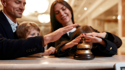 Fun time. Happy family checking in hotel at reception desk. A son and a daughter are ringing a service bell together with parents while standing at the hotel reception.