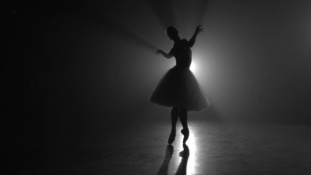 Ballet dancer in tutu performing, jumping on stage. Ballerina practices on floor in dark studio with smoke. Slow motion.