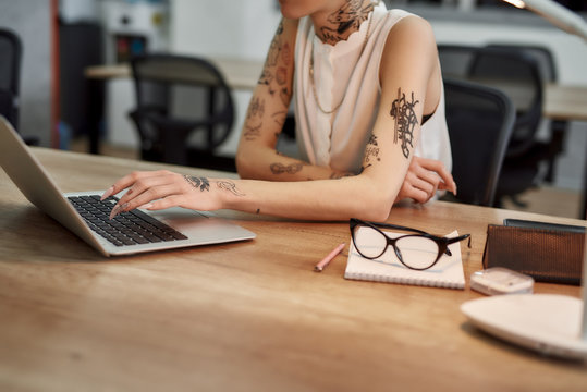 Checking email. Cropped photo of young tattooed woman typing something on the laptop while sitting at her working place in the modern office