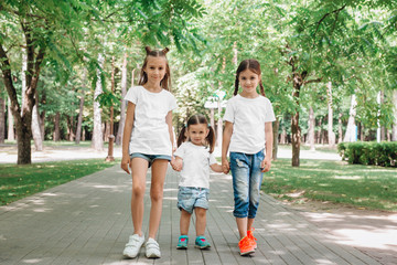 Three little sisters in white t-shirts stand holding hands in park outdoor. Mock up.