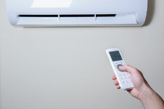 Close Up Shot Of Hand Holding The Remote Control Of Newly Installed White Air Conditioner. Working AC Hanging On The Pastel Color Wall With A Lot Of Copy Space For Text. Background.