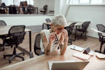 Feeling frustrated. Tired young tattooed business woman keeping eyes closed, holding her glasses in...