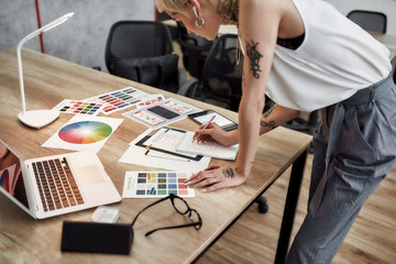 Creative female. Young blonde tattooed female designer making some sketches while standing near office desk. Working with color swatch samples