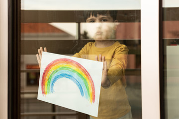 Little kid holding a drawing of a rainbow through the window