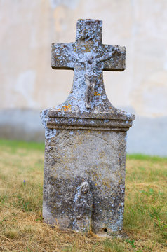 Ancient aged grave-stone with crucifix afore medieval wall. Old gravestone with holy rood in ancient cemetery. Ab olim tombstone in front of wall of old church. Tomb stone on grass in old cemetery.