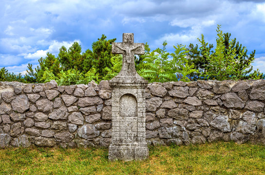 Ancient aged grave-stone with crucifix afore medieval wall. Old gravestone with holy rood in ancient cemetery. Ab olim tombstone in front of hard rock fence. Tomb stone on grass in old cemetery.