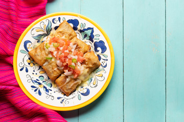 Mexican chimichanga with melted cheese and pico de gallo sauce on turquoise background