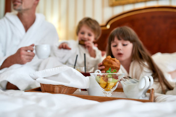 Obraz na płótnie Canvas Healthy and tasty. Family in white bathrobes having breakfast in bed, eating pastries and drinking coffee in luxurious hotel room. Focus on fruit cup on the tray. Food, resort, room service concept