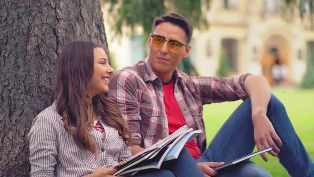 Young students sitting outdoors under the tree of campus park and having converation. Two young classmates talking sitting on a green grass after or betwin classes.