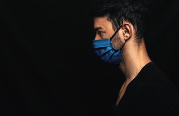 Side view image of Caucasianman wearing medical blue mask on the face during virus pandemic lockdown posing on the black wall with copy space. Male with disposable face mask to prevent the coronavirus