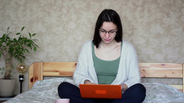 Young attractive girl is actively browsing the news feed on a laptop while sitting on a bed at home. Young Cute girl is working on a laptop while sitting on a bed.