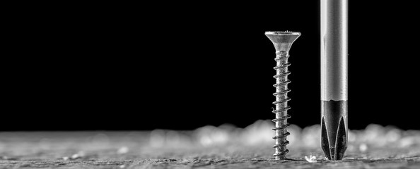 The screw and screwdriver close up on black background. Joinery and construction work. Monochrome photo. Panoramic banner. - obrazy, fototapety, plakaty