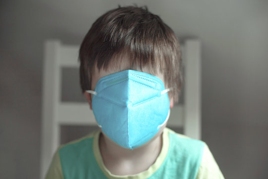 A little boy, a European child in a blue medical mask on his whole face sits on a chair. Self isolation. Panic, fear of the virus, fear of disease.
