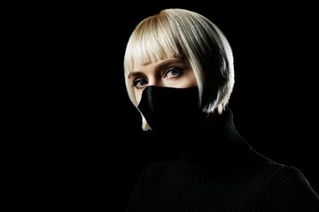 Woman wearing protective handmade black face mask looks at camera, posing in the darkness. Copy, empty space for text