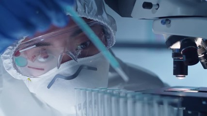 Asian female virologist in protective suit, mask, glasses and gloves dripping blue liquid from pipette into test tubes while working on antiviral vaccine at desk with microscope on it in laboratory - Powered by Adobe