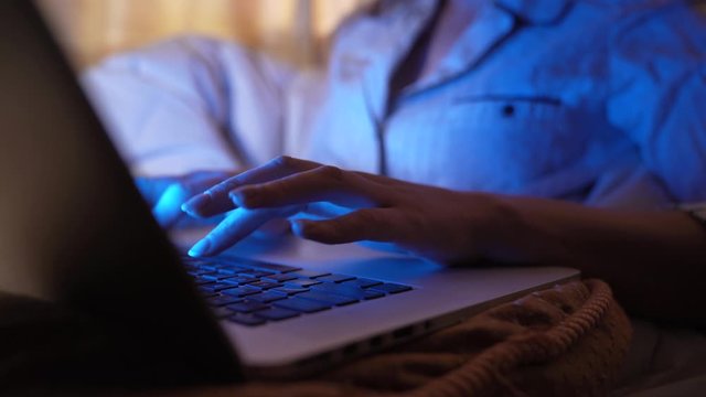 This close up video shows the hands of someone in pajamas working at home and typing on a laptop with the computer screen glowing.