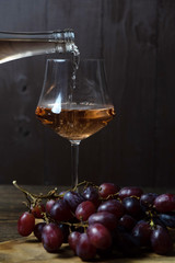 Rose wine is poured into a glass goblet from a bottle. In the foreground, a bunch of grapes is out of focus. The dark key. Vertical orientation
