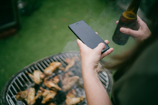 Woman taking a photo with the mobile to a grill of chicken wings cooking on the barbecue while having a beer in the backyard.