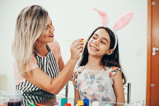 A mother and daughter celebrating Easter, painting eggs with brush. Happy family smiling and laughing, drawing on face