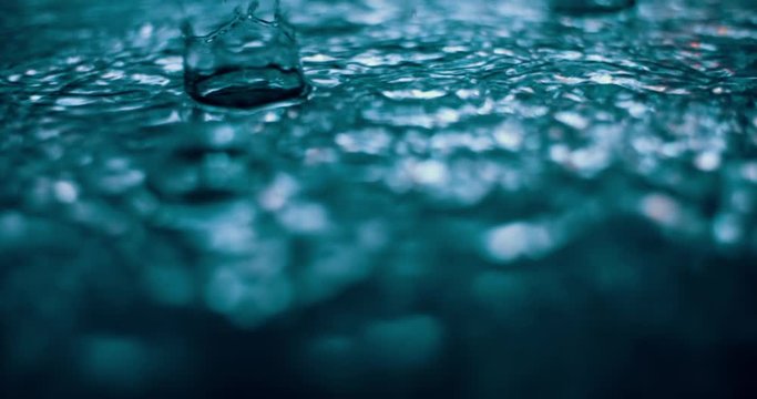 Autumn raindrops falling into puddle on asphalt at night. Big city lights mirror reflection on water surface. Close up of raining outside in slow motion. Rainy evening in street. Slow motion shot