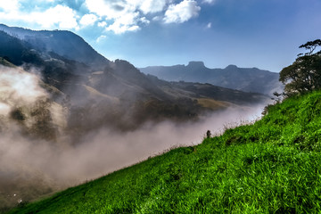 Imagem de vale montanhoso com nuvens, durante o amanhecer e silhueta da Pedra do Baú ao fundo, no...