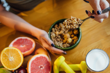 Cropped image, woman eating corn flakes cereal, fresh oranges and dumbbells while sitting and having breakfast at the kitchen table