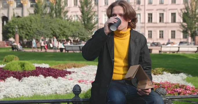 The young man drinks coffee and eats sweet cake in the park, he smiles, is dressed in a yellow sweater and black raincoat or jacket, Jeans trousers, flowers and green grass on background, sunny day