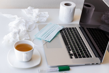 A laptop with a white cup of tea and saucer on quarantine, a medical mask on the keyboard, a roll of toilet paper and used napkins,  thermometer. Everything on a white table.