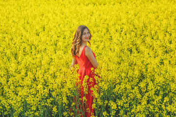Fototapeta premium Young beautiful girl in a red dress close up in the middle of the yellow field with the radish flowers