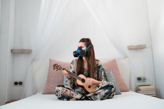  Young white woman in antibacterial mask and floral dress playing her ukulele sitting on the bed at home due to the isolation of COVID-19. - Powered by Adobe