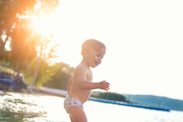 Cute adorable cheerful caucasian little blond toddler boy enjoy having fun playing at lake or river beach water on warm sunset evening time outdoors.. Happy childhood vacation at countryside concept