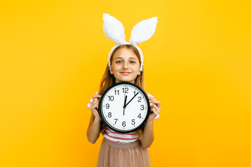 Happy beautiful girl on her head with rabbit ears, with a wall clock in her hands on a yellow...