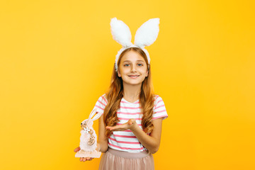 Obraz na płótnie Canvas A happy girl on a head with rabbit ears holds a wooden Easter Bunny on a yellow background