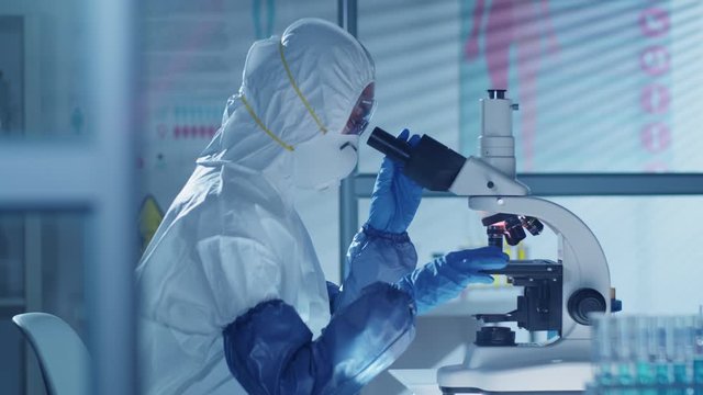 Side view of unrecognizable virologist in protective mask, suit and gloves looking through microscope while researching on Covid-19 treatment in laboratory with biohazard sign