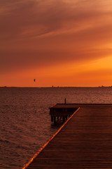 Wooden pier on a warm light during sunset, with orange sky and a Kite surf in the background