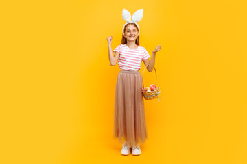 Full length, a small attractive girl on a head with rabbit ears, holding a basket of Easter eggs and showing a victory gesture