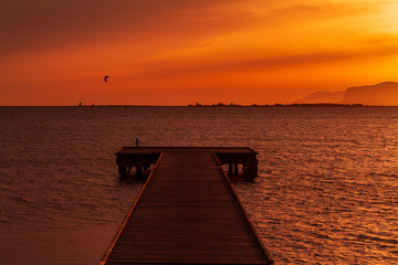 Fototapeta na wymiar Wooden pier on a warm light during sunset, with orange sky and a Kite surf and two mountains silhouette in the background