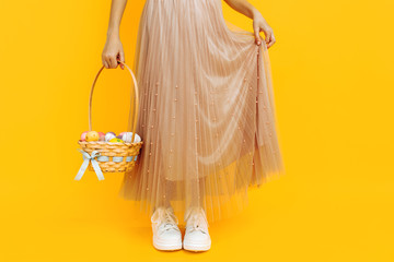Close-up photo of legs, a girl holding a basket with Easter eggs on a yellow background