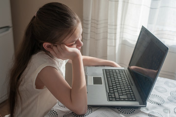 A school-age girl sits at home at a table and studies using a laptop. Distance Education, Quarantine