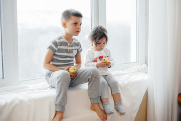 Brother and sister are sitting on the windowsill playing and eating apples. Happiness