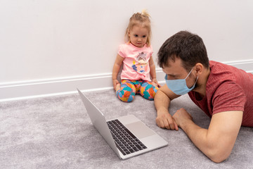 Father in facial mask tries to work at home at the period of quarantine because coronavirus,his daughter sit near him,looks at screen of laptop. Concept of home office work with family. Copy space.