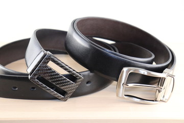 Two beautiful, bilateral belts, made of smooth black leather and carbon, steel fittings, lie on top of each other, on a table made of light wood. Expensive gift option for men, women, premium leather,