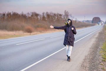 COVID-19. A girl in dark clothes and a gas mask outside the city on a cloudy day stands on the edge of the road.