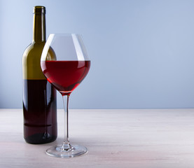 Low angle perspective of a crystal clear glass with a traditional glass filled with dark red wine and bottle and a thin leg on a wooden table with a light gray background with copy space for your text
