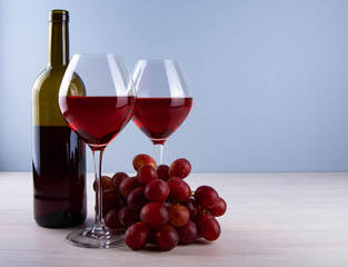 Low angle perspective with a two traditional glass filled with dark red wine and red grape and bootle and a thin leg on a wooden table with a light gray background with copy space for your text